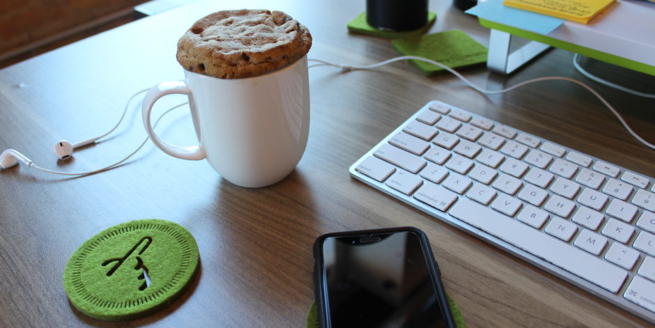 A cookie resting on a coffee mug next to a smartphone and a computer keyboard, meant to show the connection or competition between technology and mental health. 