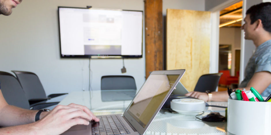 A man on the left taking notes on his laptop while another man on the right is looking at an interface on a television for a usability test