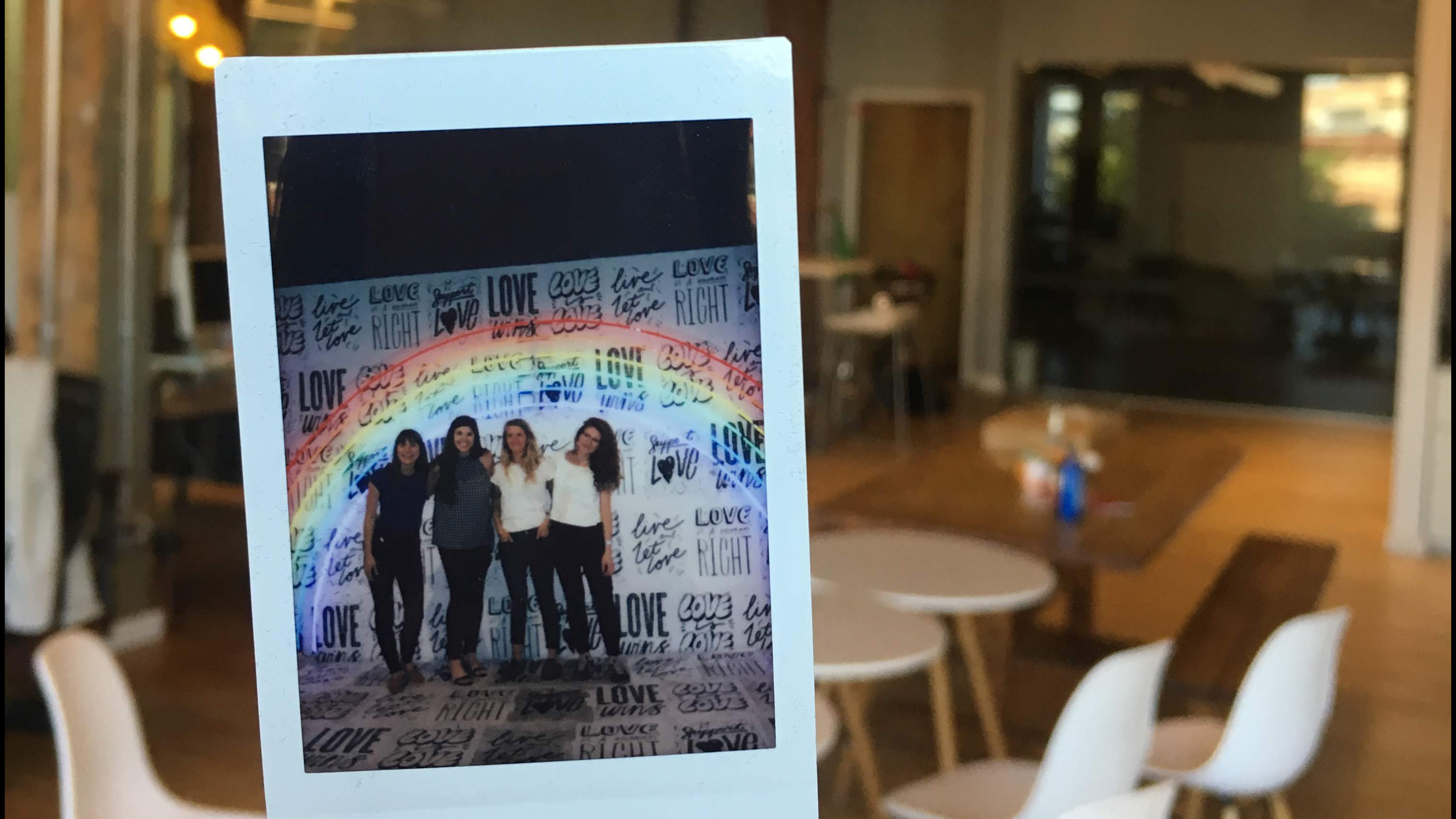 Polaroid of four women against a background that has several phrases about love written on it and a large neon rainbow overhead