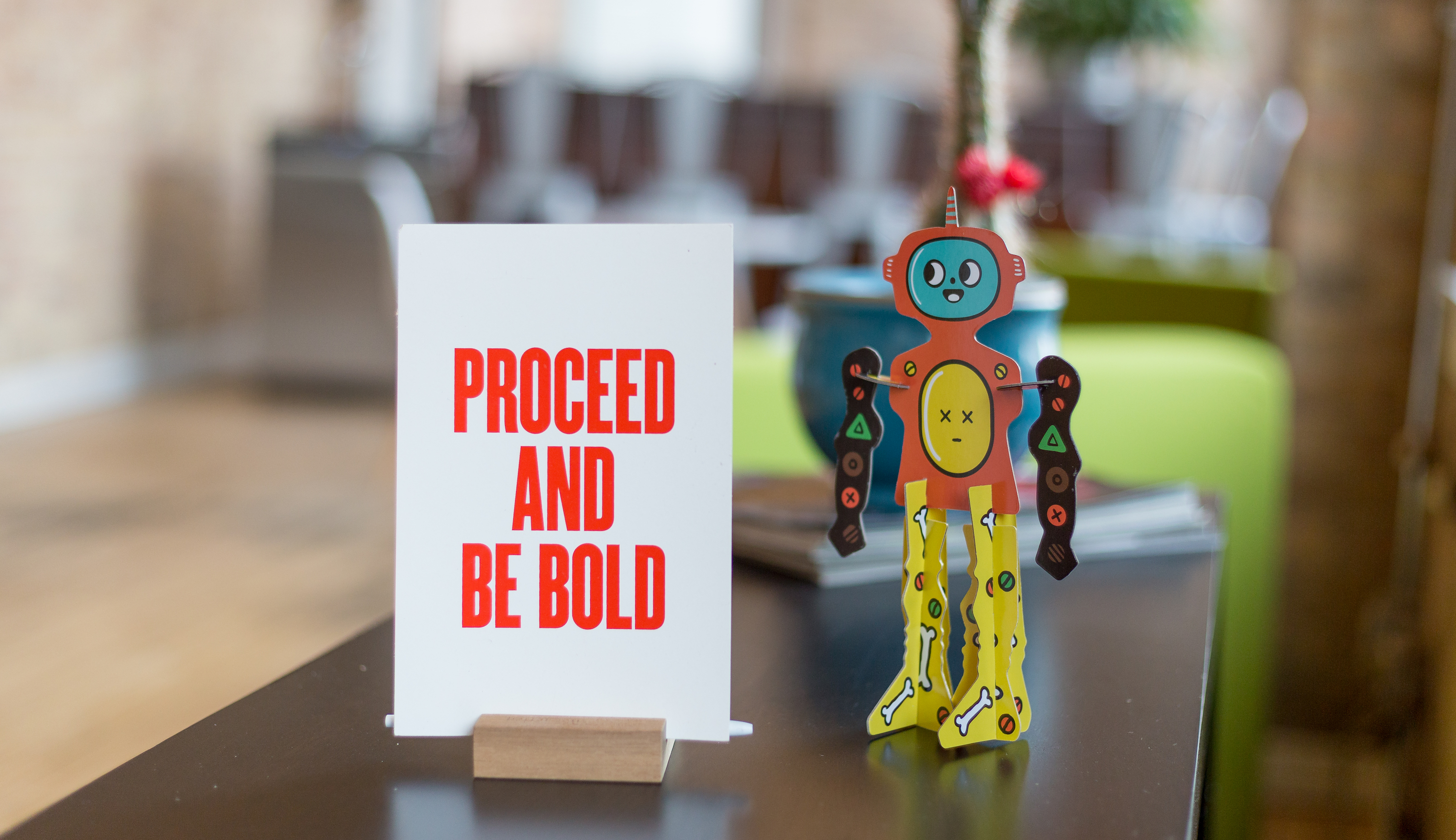 A white card with red letters reading "Proceed and be bold" next to a cardboard cutout robot figurine