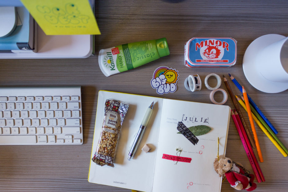 A photo of a desk taken from the top down. The desk has an open notebook with doodles, leaves, colored pencils, a cup of coffee, and other assorted knick-knacks