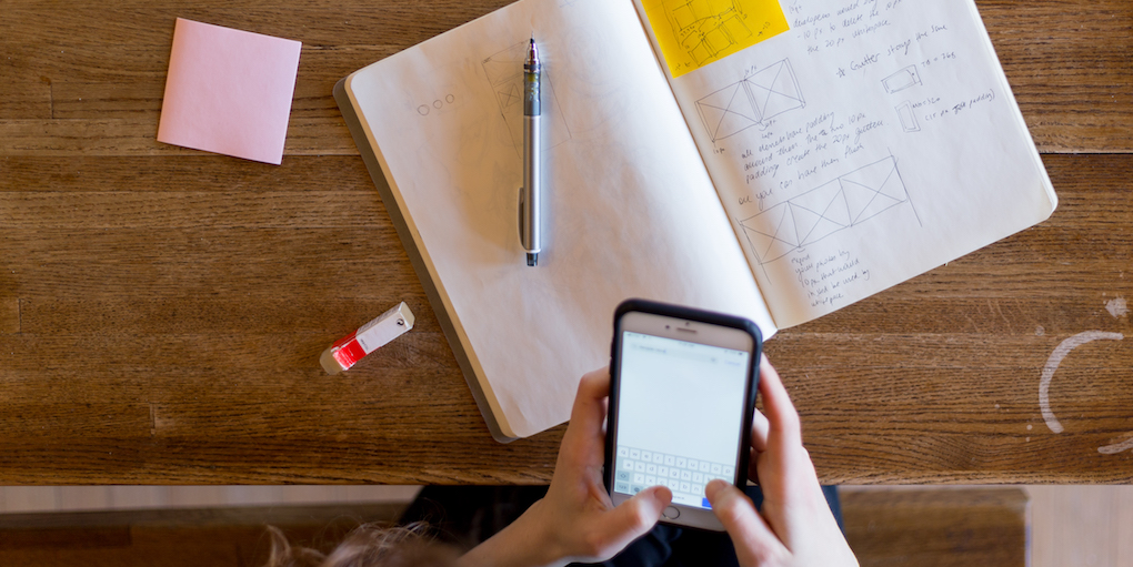 A photo of a desk from above with a woman holding an iPhone, a notebook full of sketches, a Post-It note, a pencil, and an eraser