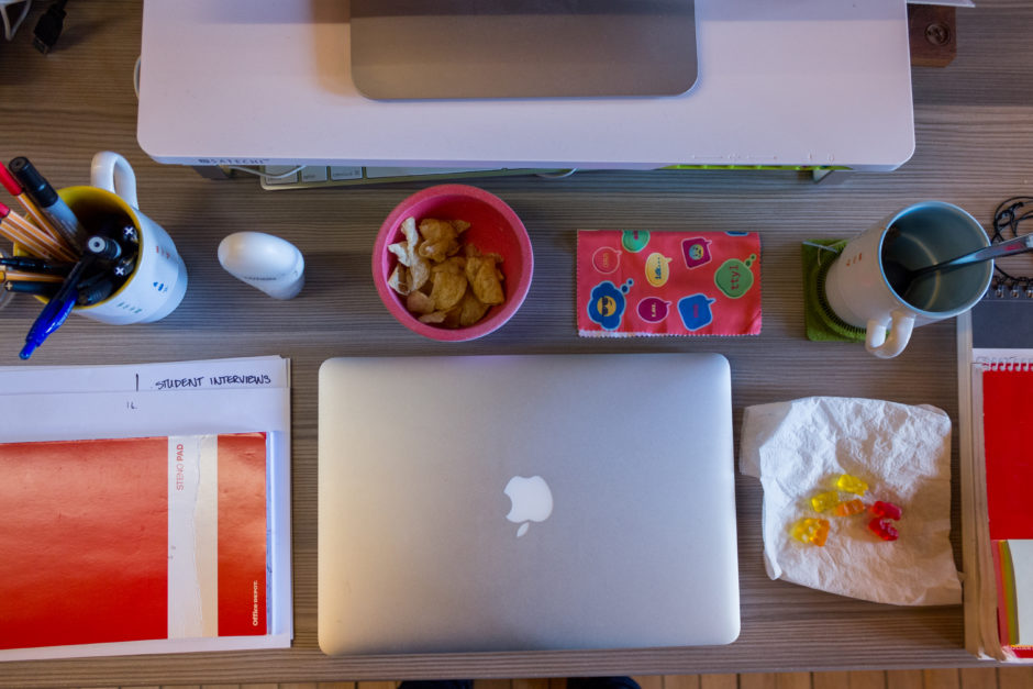 A photo of a desk taken from the top down. The desk has a closed Macbook, a coffee cup full of pens and pencils, a small bowl of snacks, an open notebook, and an empty coffee cup.