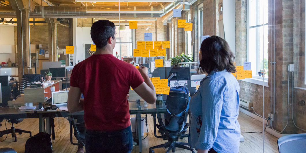 Two Fuzzy Math UX researchers working at a glass wall with sticky notes on it. One person is talking and gesturing while the other is writing on a sticky note on the glass wall.