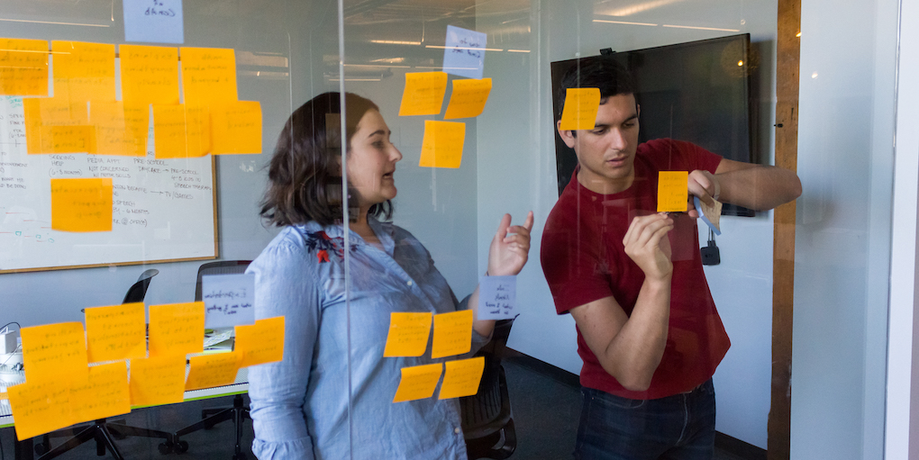 Two Fuzzy Math UX researchers working at a glass wall with sticky notes on it. One person is talking and gesturing while the other is writing on a sticky note on the glass wall.