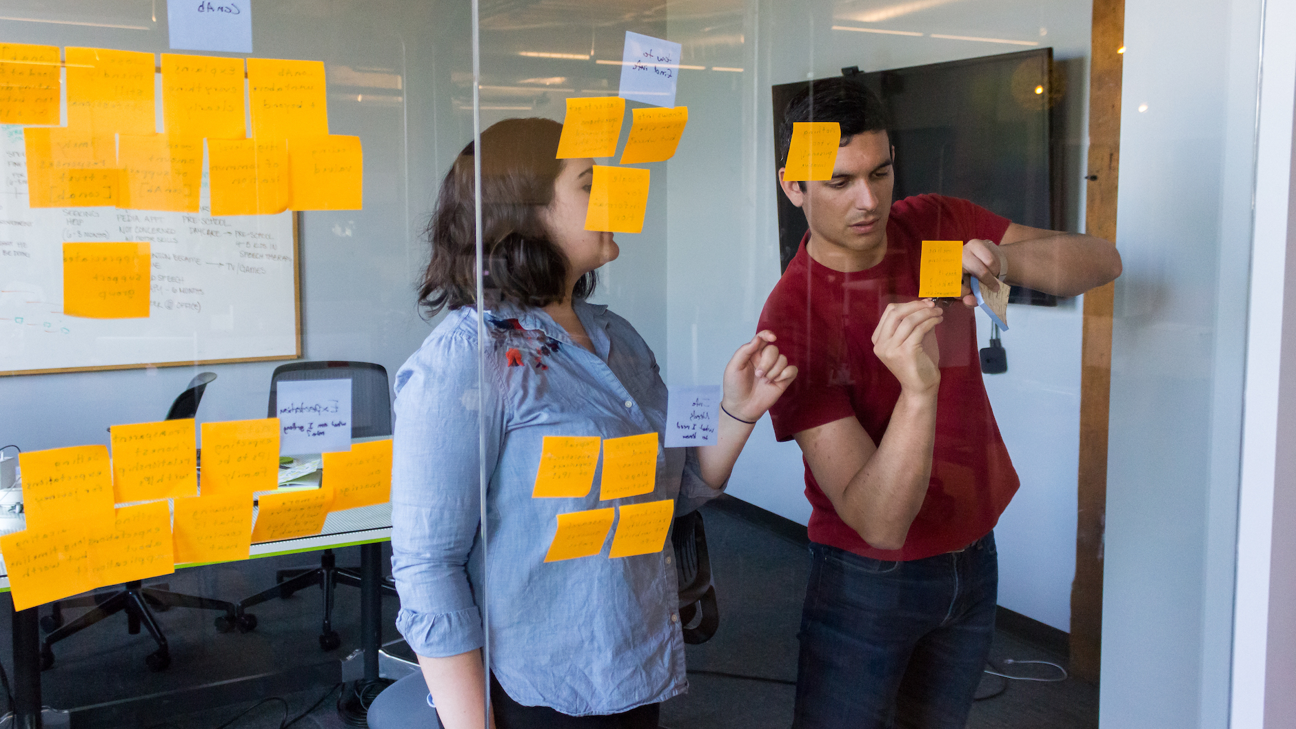 Two Fuzzy Math UX researchers are conducting synthesis of user experience testing research, working at a glass wall with sticky notes on it. One person is talking and gesturing while the other is writing on a sticky note on the glass wall