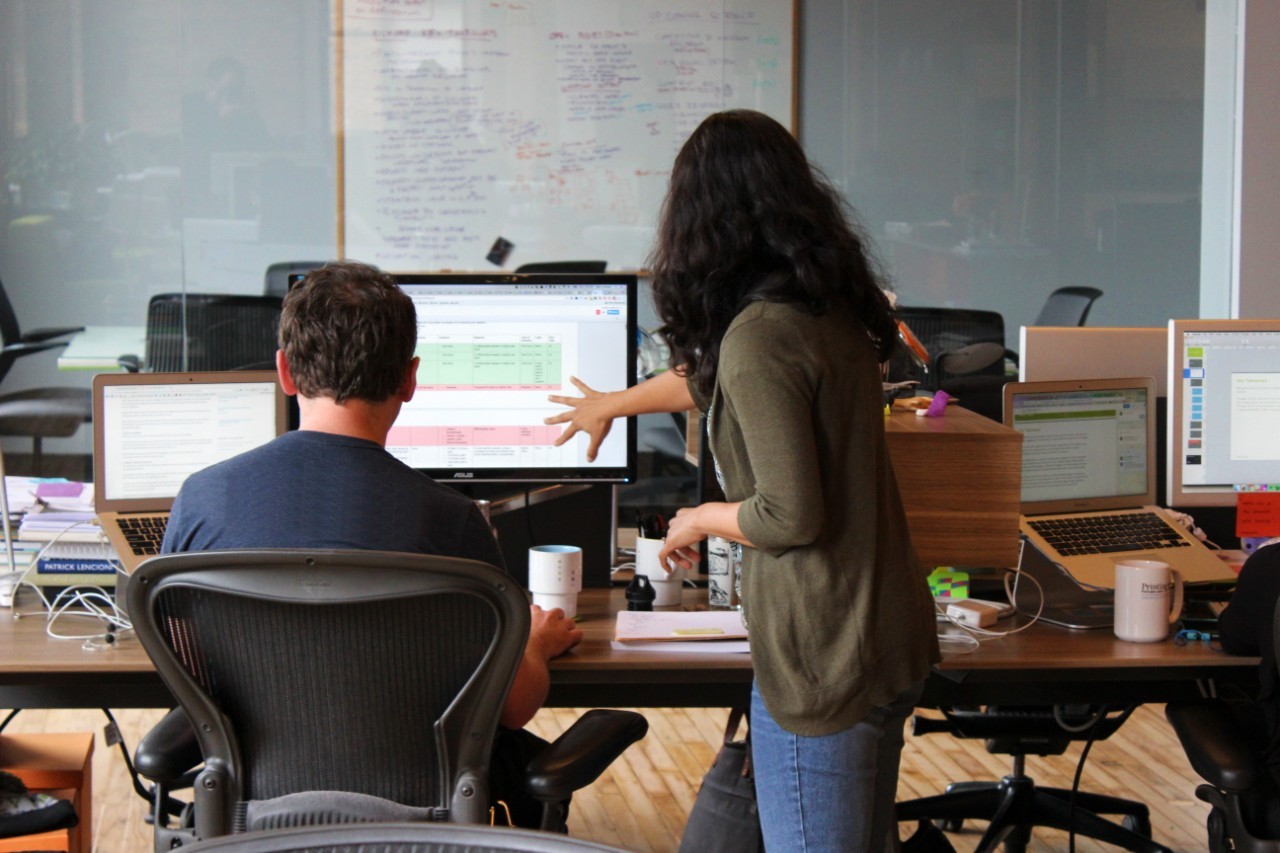 Two UX designers evaluating the end user experience of a product and looking at a computer screen. One is sitting down at the computer, and the other is standing, gesturing to the screen.