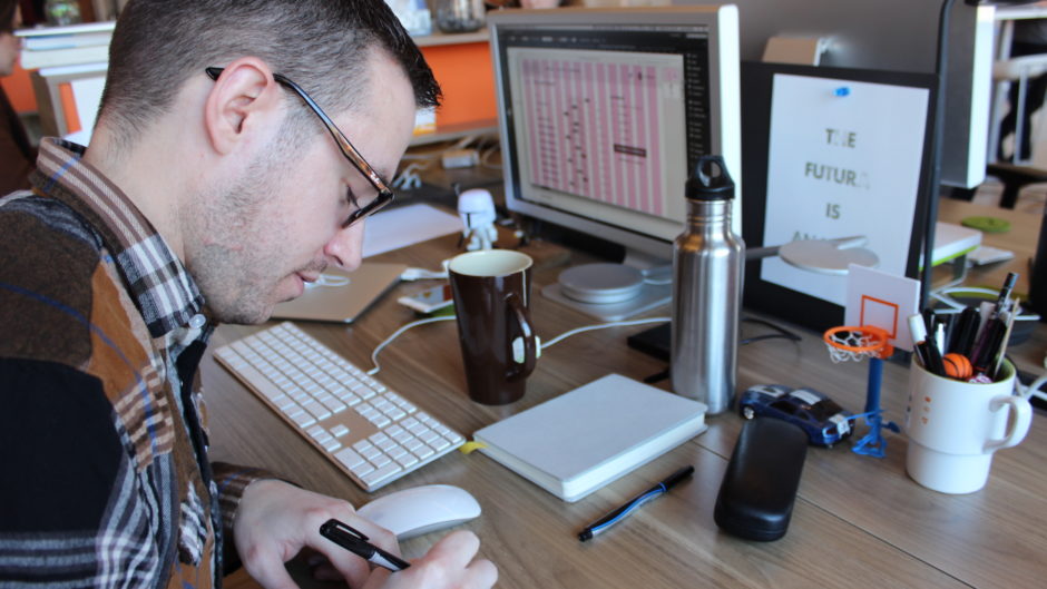 Depending on where the project is in its lifespan, a day in the life of a UX designer often includes wireframing. A UX designer sits at a desk, drawing in a notebook. The computer in the background shows a wireframe in progress.