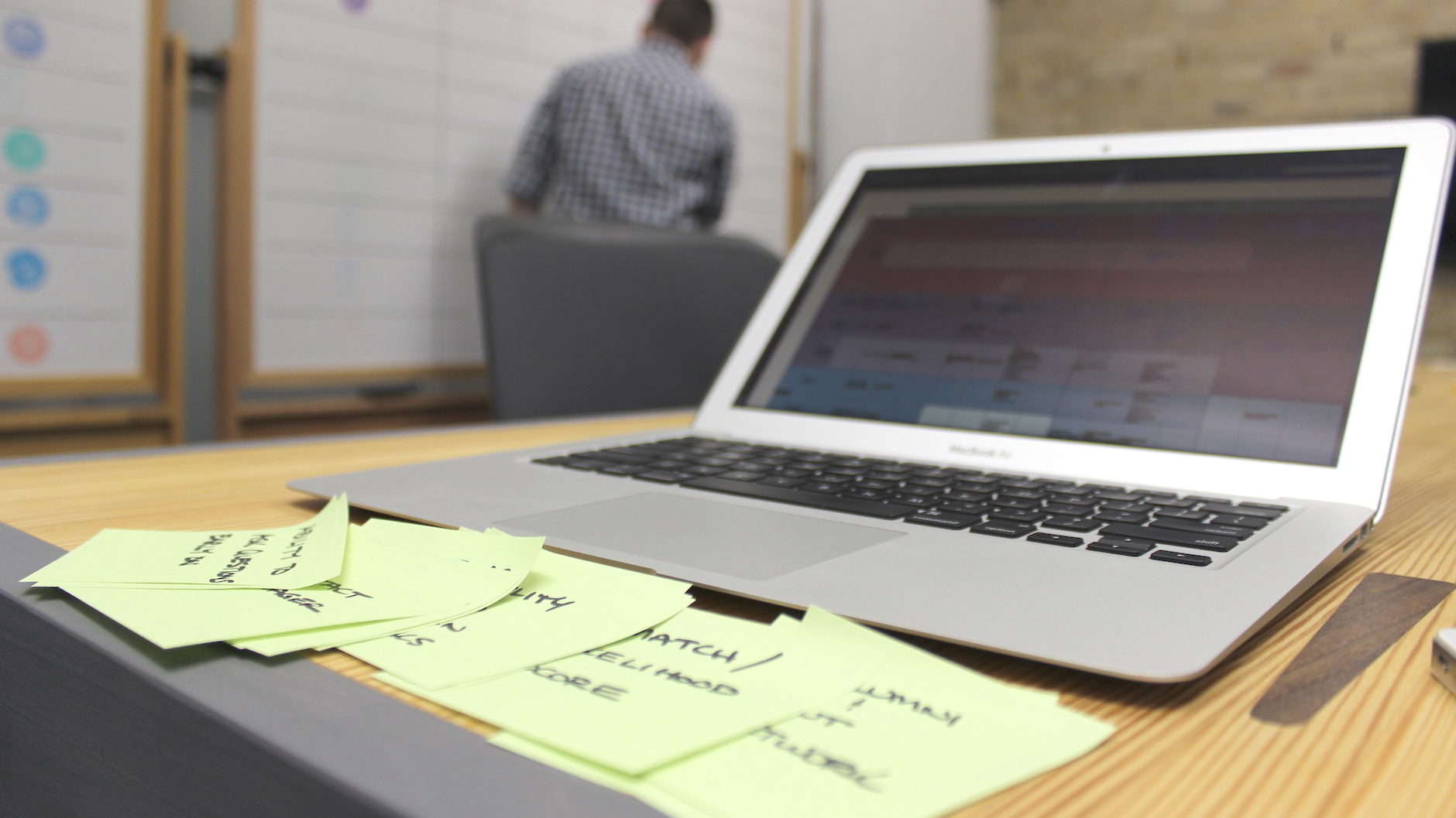 A laptop sitting on a desk. There are light green sticky notes on the desk describing the difference between user experience and usability. There is a UX designer writing on a whiteboard in the background.