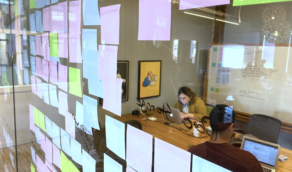 Post-it notes on glass wall looking into a conference room where researchers are synthesizing enterprise ux research. 