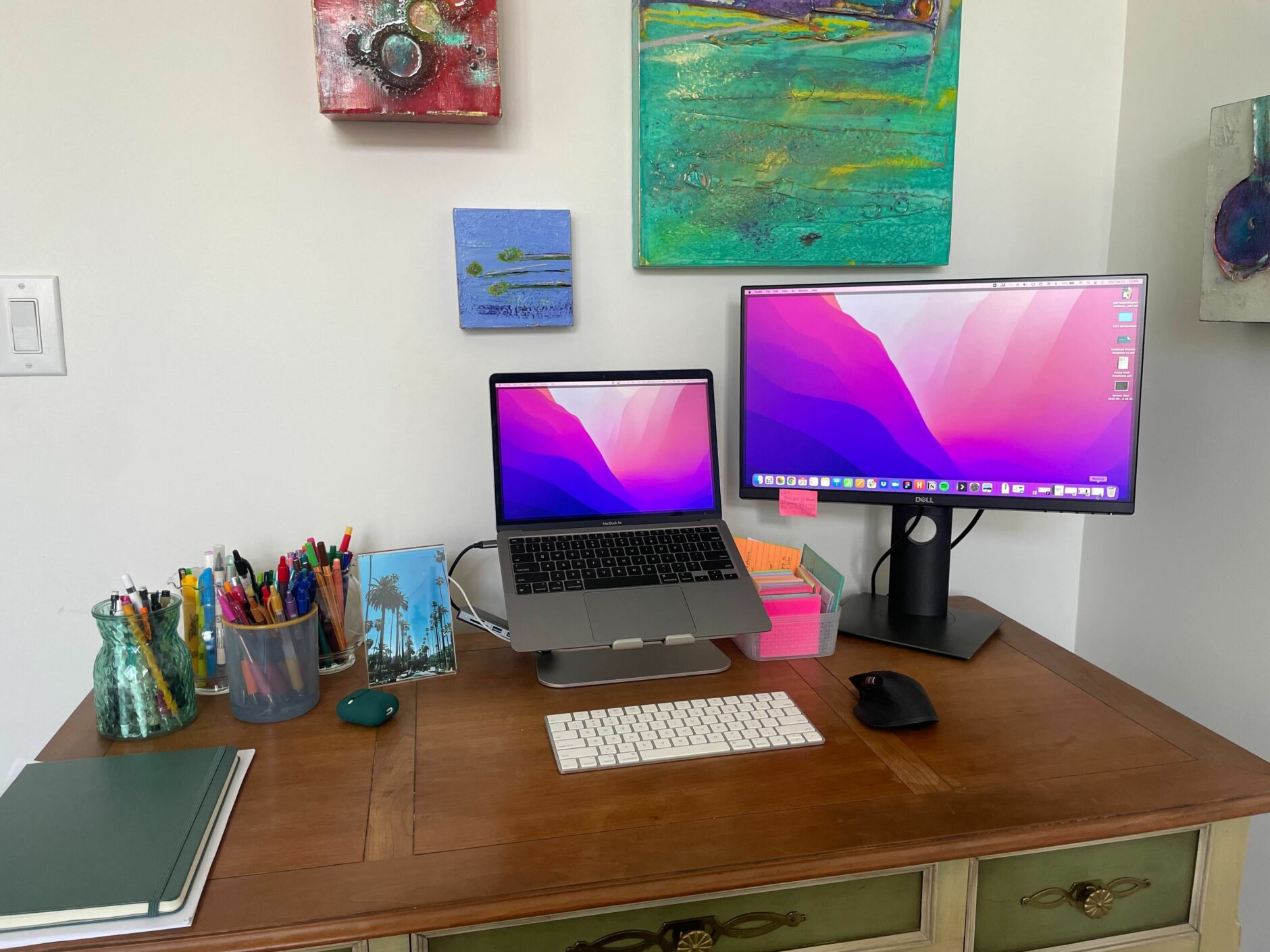 Desk setup with a monitor and laptop. There is artwork in the background and multiple colored pencils in cups on the desk.