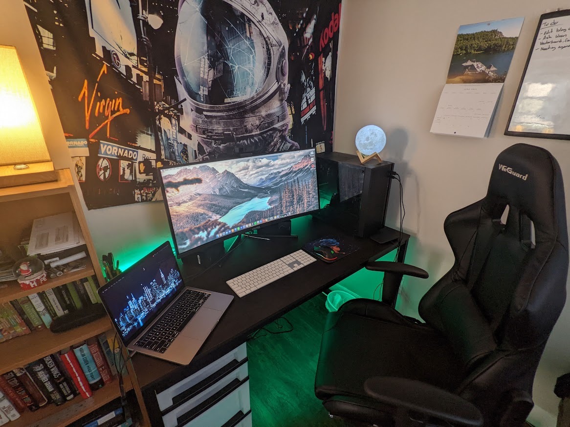 Desk setup showing a central monitor with a laptop on the side. Desk has green background lighting and a moon lamp. A tapestry with an astronaut in NYC is in the background. 