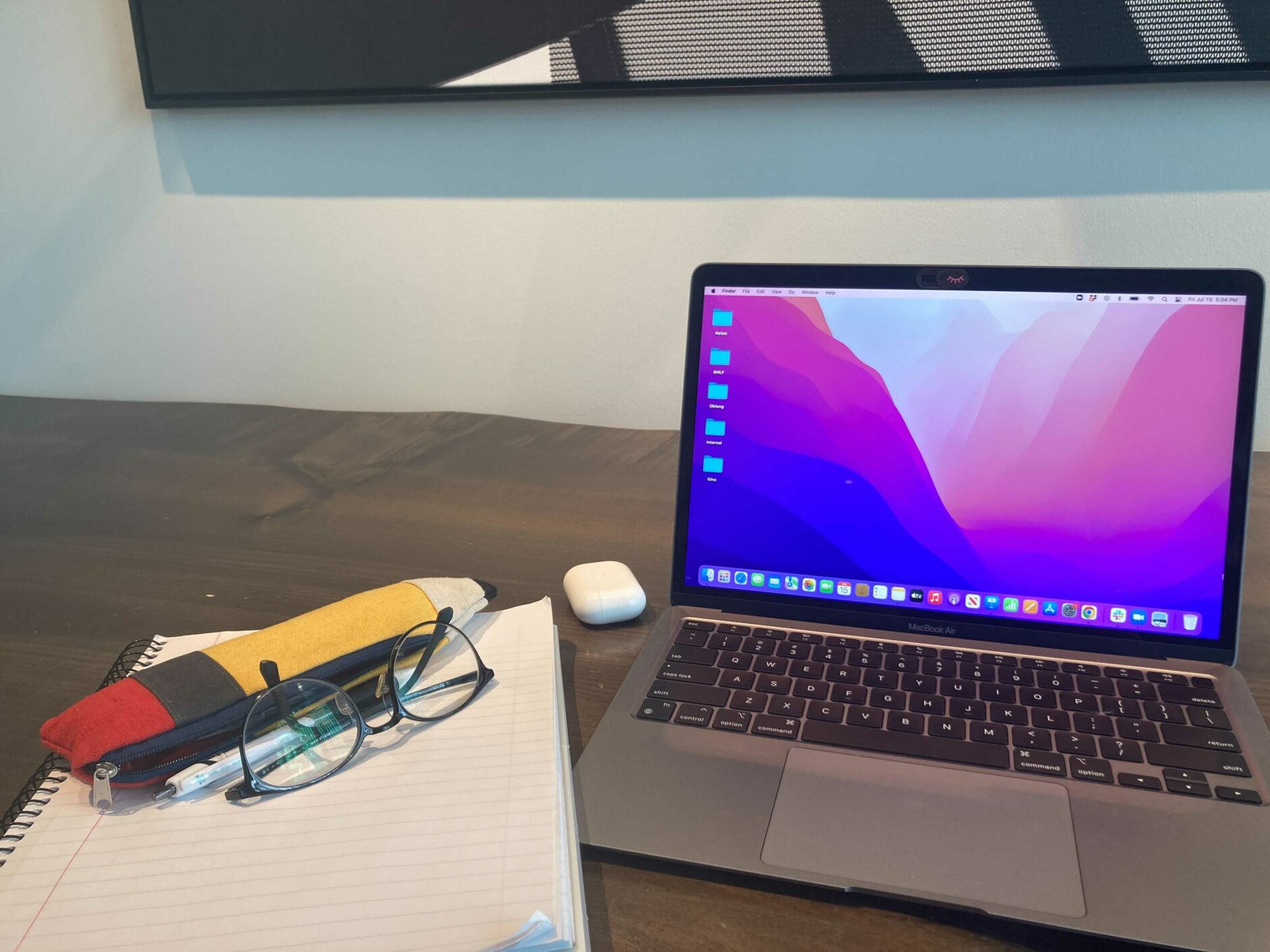 Desk setup with a laptop monitor and a variety of desk items like airpods, glasses, and a pencil shaped pencil case.