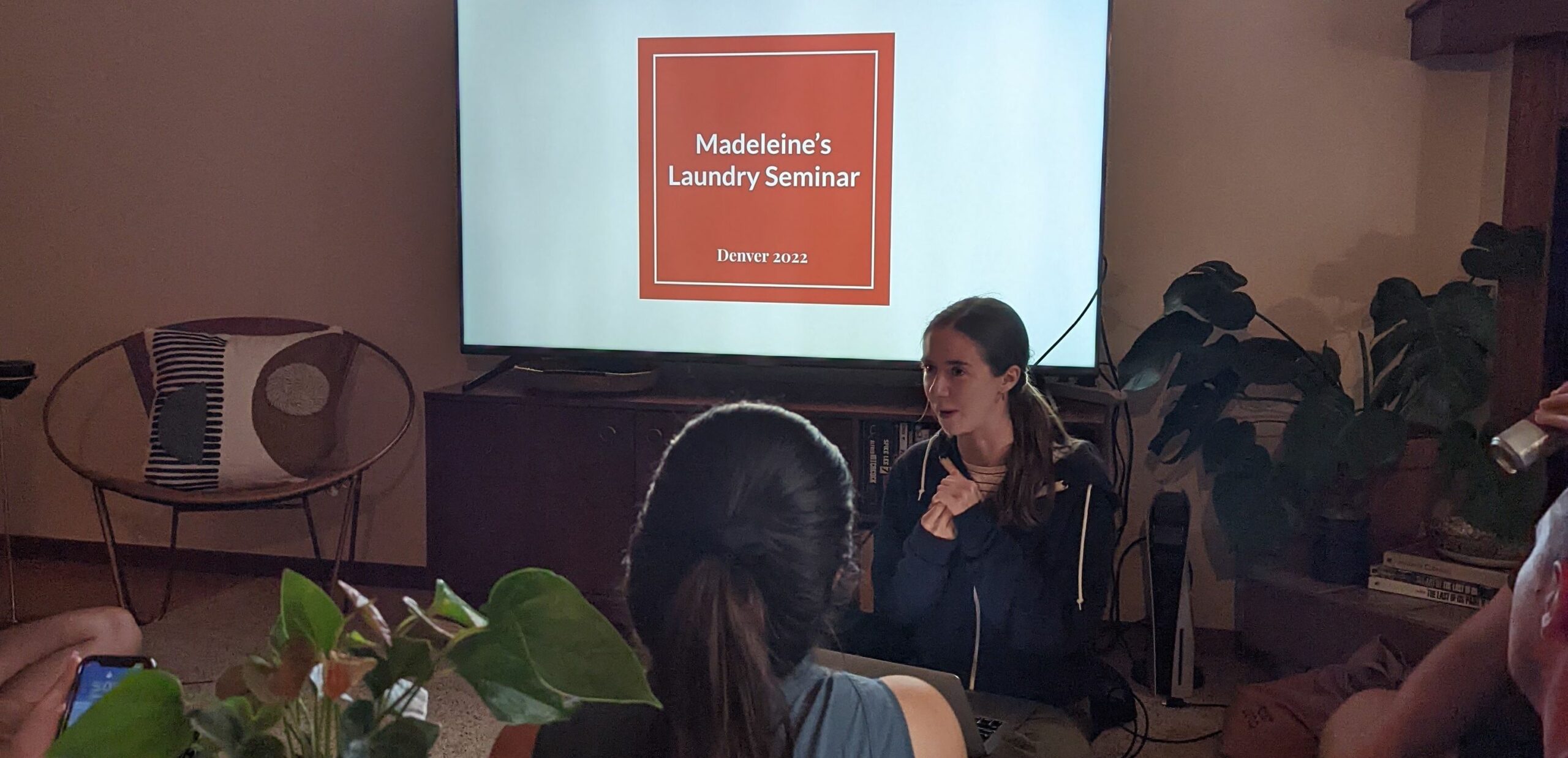 Picture of Madeleine sitting in front of a TV showing a slide projection titled 'Madeleine's Laundry Seminar'